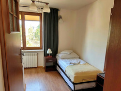 na zdjęciu prezentowany jest pokój jednoosobowy, wyposażony w łóżko ubrane w pościel, na którym leży ręcznik, pod oknem usytuowane jest biurko z telewizorem i lodówką