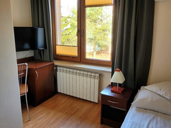 na zdjęciu prezentowany jest pokój jednoosobowy, wyposażony w łóżko ubrane w pościel, na którym leży ręcznik, pod oknem usytuowane jest biurko z telewizorem i lodówką